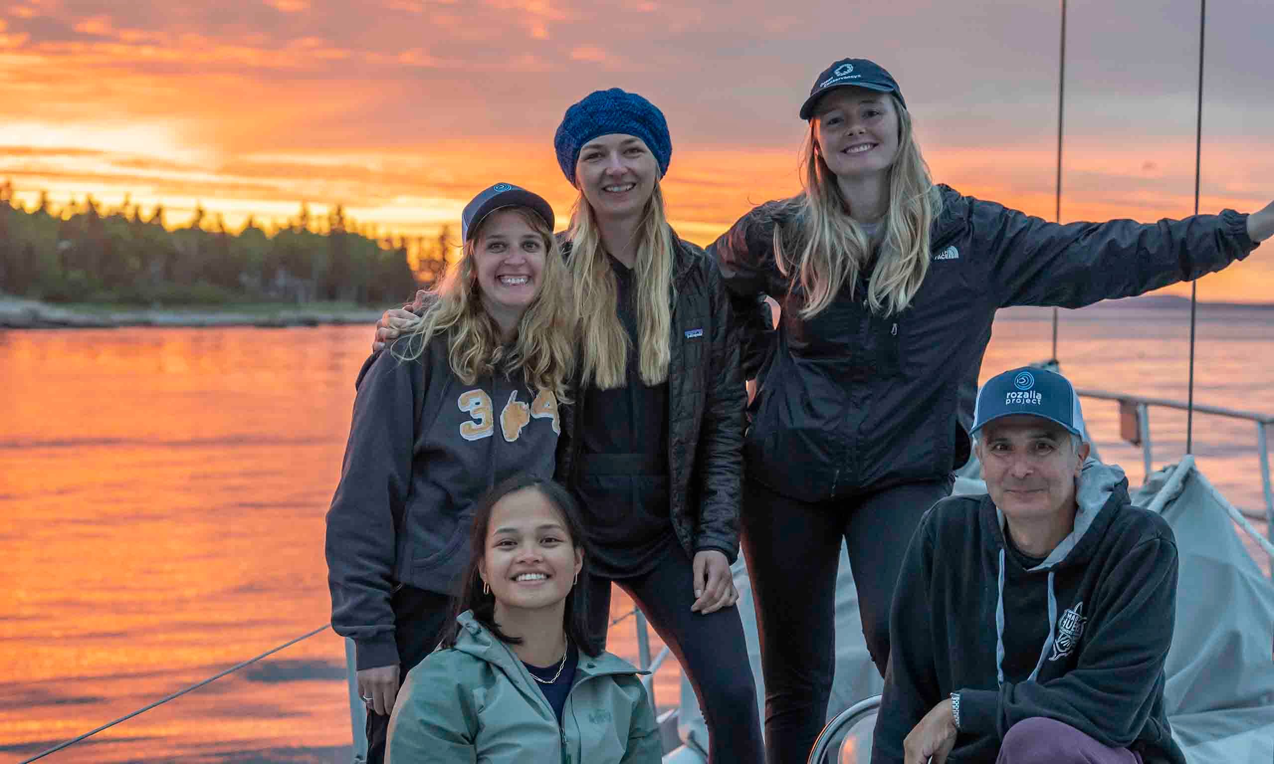 A group of Ocean Conservancy staff smiling aboard the American Promise sailboat with their backs to an incredible orange and purple sunset.