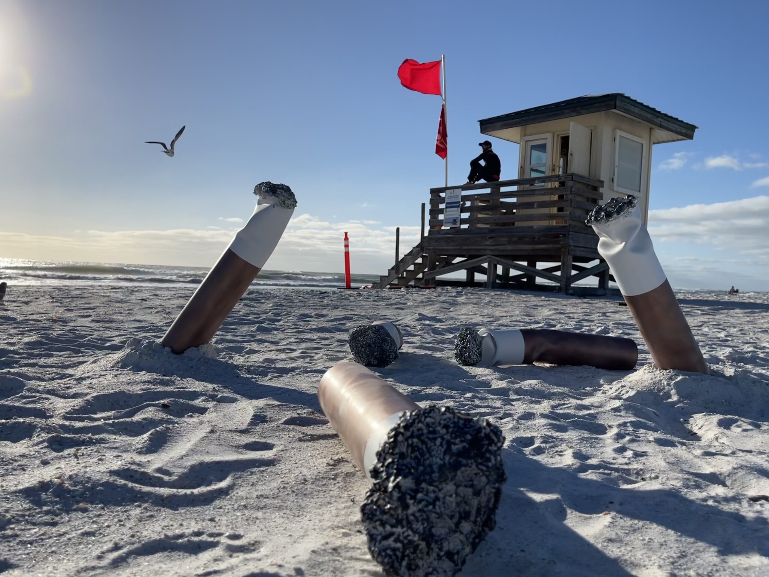 Giant cigarette butts on a beach with a lifeguard tower in the background.