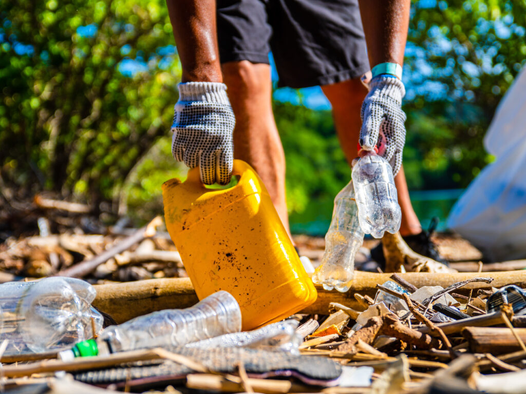 An up close view of gloved hands picking plastic bits off the beach. A yellow plastic container is being lifted by gloved hands off a beach shoreline.