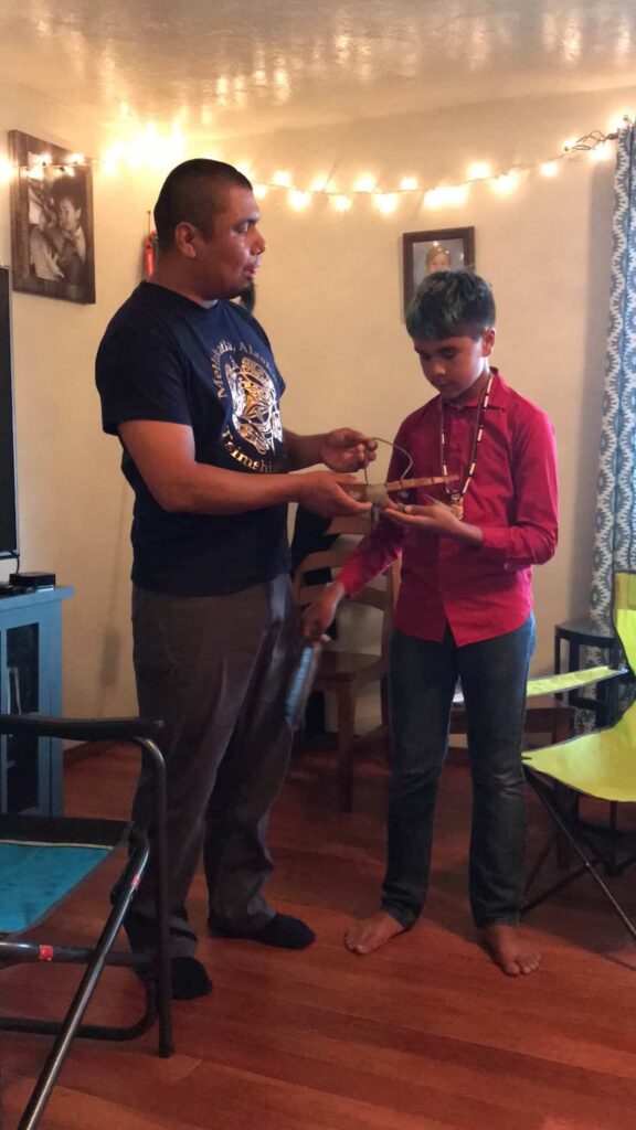 Halibut hook: K’alaagáa Íihlangaa (Nathaniel) Blake receiving a Halibut Hook and listening to Tlingit teachings from his uncle Xeetli.éesh (Lyle) James. Xeetli.éesh was one of many leaders who stood to acknowledge this moment and gift knowledge and tools to aid K’alaagáa Íihlangaa as he grows in the responsibility to care and provide for his community.