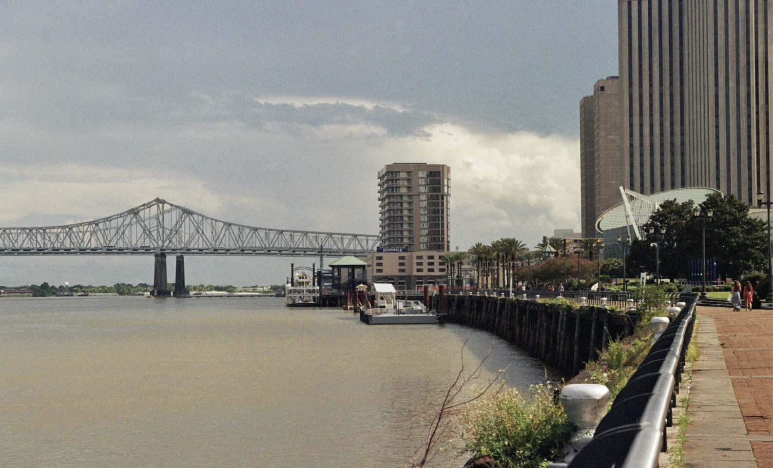 View of the Mississippi River from New Orleans on an overcast day. Green foliage line the port as people walk along the shore. The the Crescent City Connector is featured.