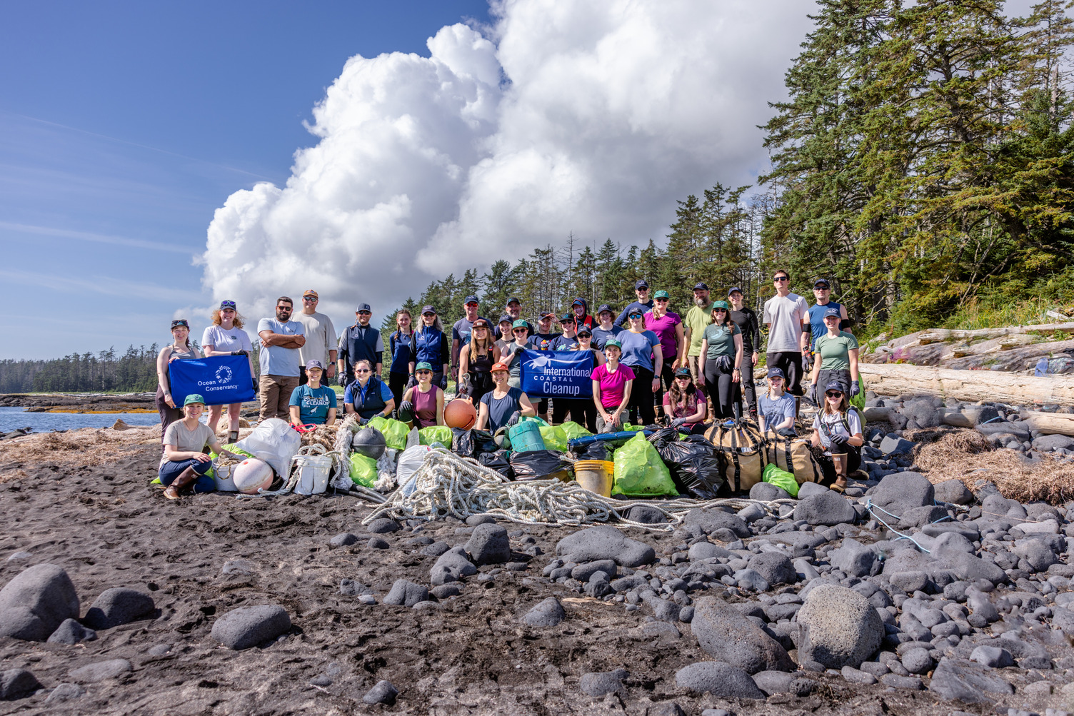 A group of volunteers stand on a rocky beach on a sunny day. One volunteer holds up an Ocean Conservancy banner.
