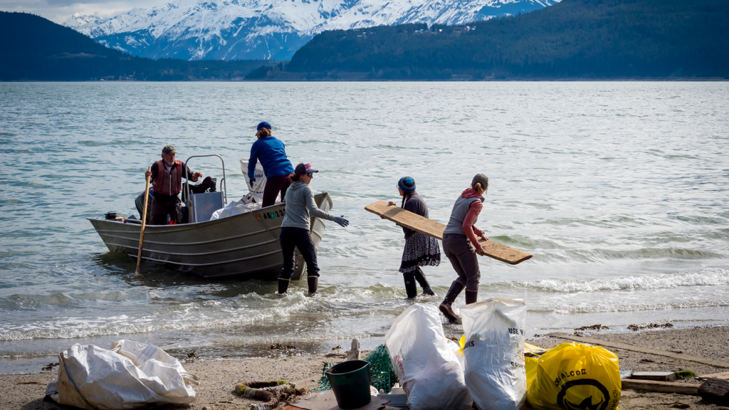 People gather on a shoreline to clean up large scale marine debris. Some people are in a row boat and some are on the shore. It seems they are removing debris by placing it in the boat.