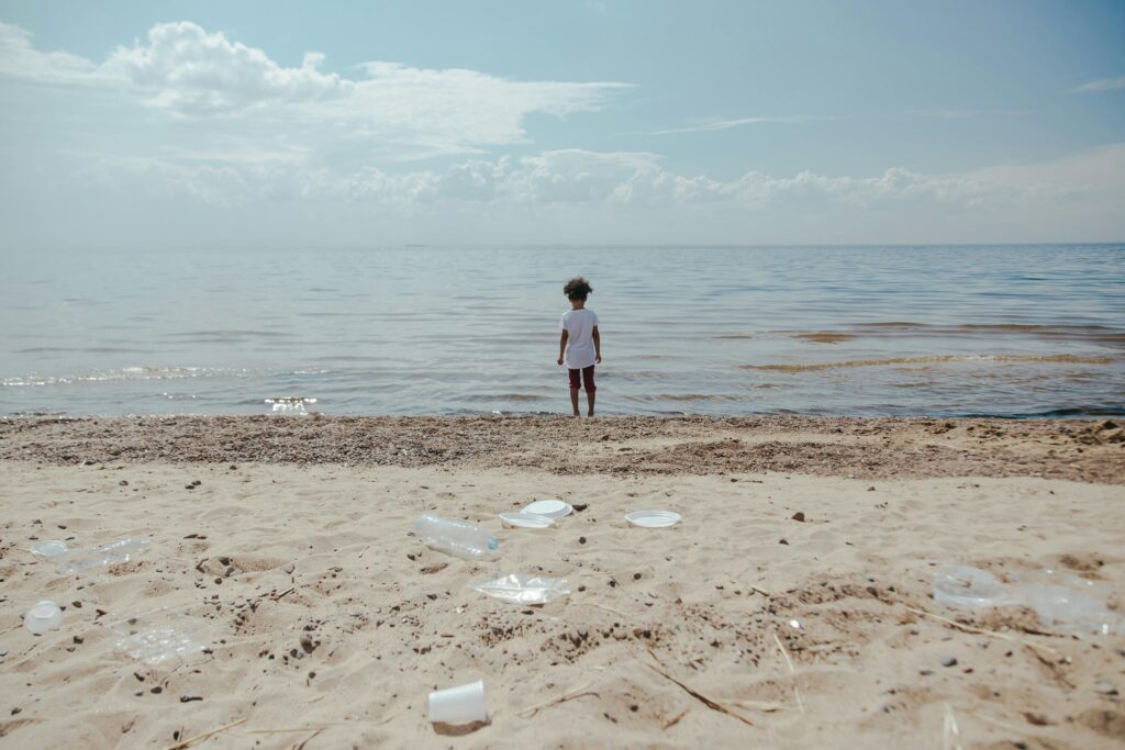 A child stands on the shore facing the ocean. In the foreground, there is plastic trash strewn about the sand.