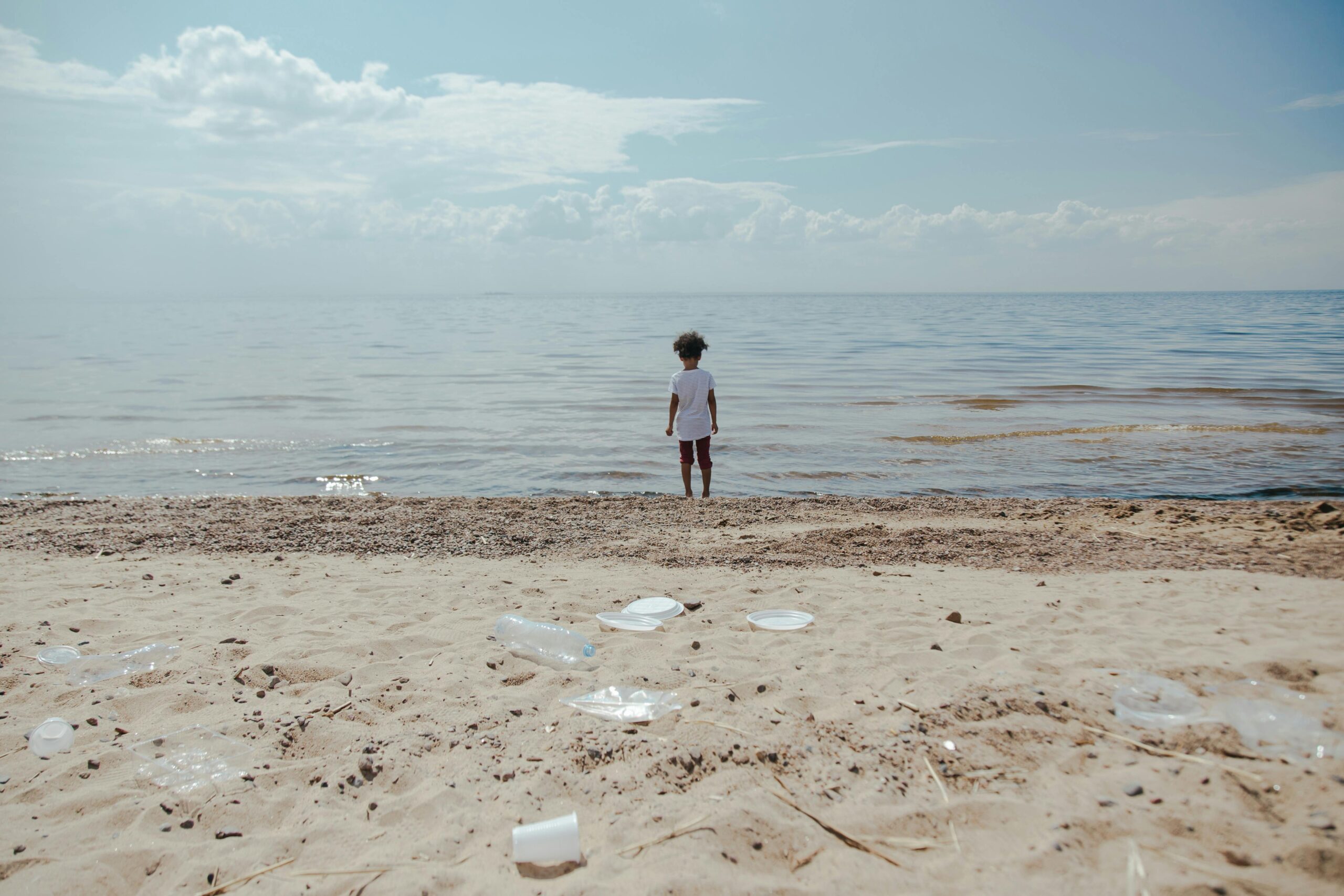 A child stands on the shore facing the ocean. In the foreground, there is plastic trash strewn about the sand.