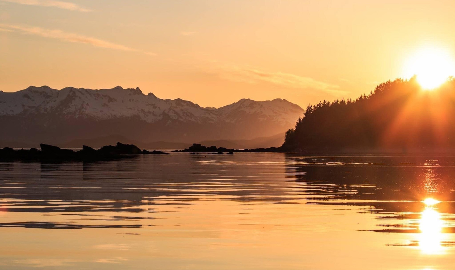 An incredible sunset in Juneau, Alaska. The sun casts an orange glow as it offers its last light peeking just above a mountain across the serene water.